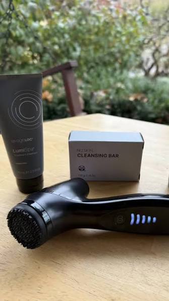 Black cleansing bar, NuSkin tube, and Black Edition LumiSpa on a wooden surface with blurred greenery in the background