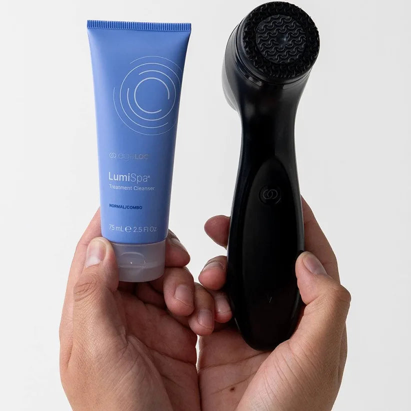 Hand holding a blue tube of LumiSpa cleanser and a black LumiSpa device on a white background