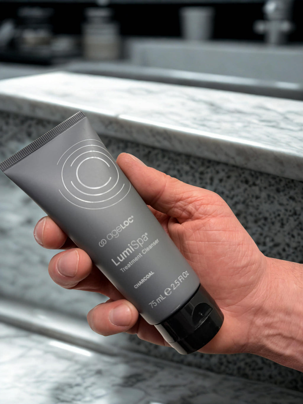 Hand holding a gray tube of LumiSpa product on a marble countertop.