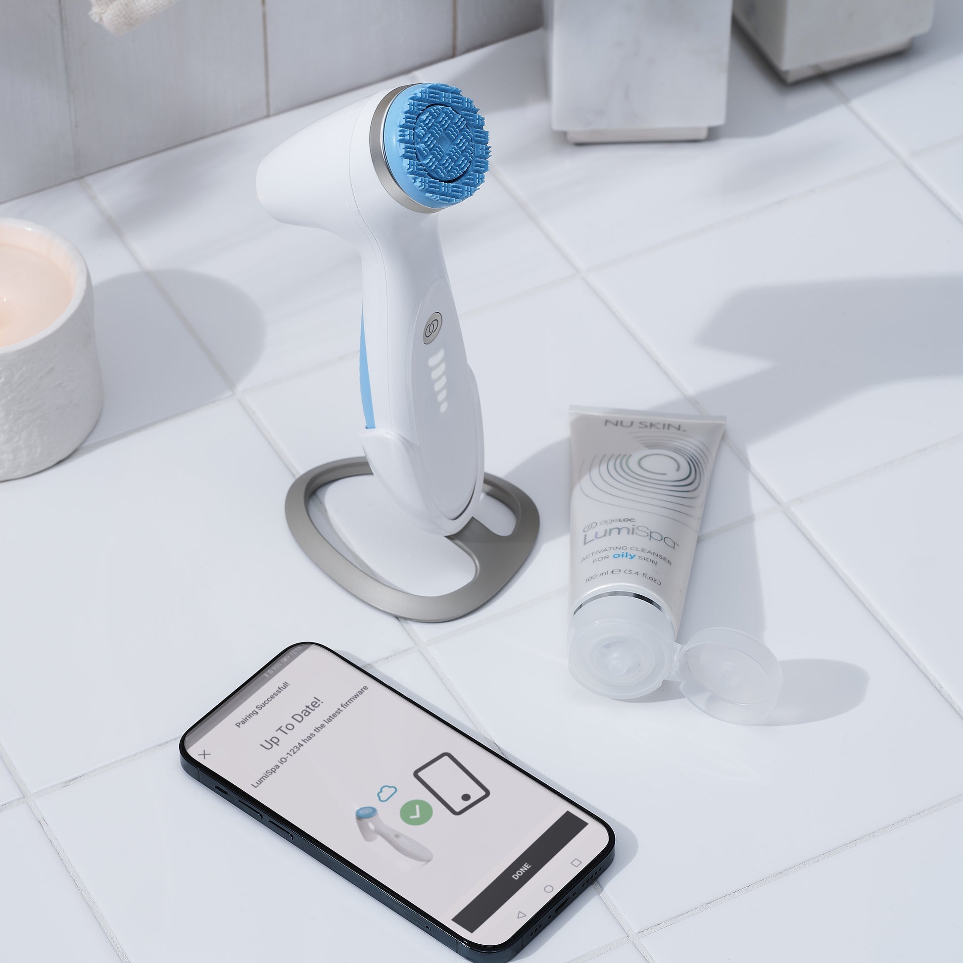 The image shows a white ageLOC LumiSpa iO skincare device with a blue accent, a magnetic charger, and a normal treatment head, displayed on a bathroom counter alongside a smartphone with a skincare app on the screen.